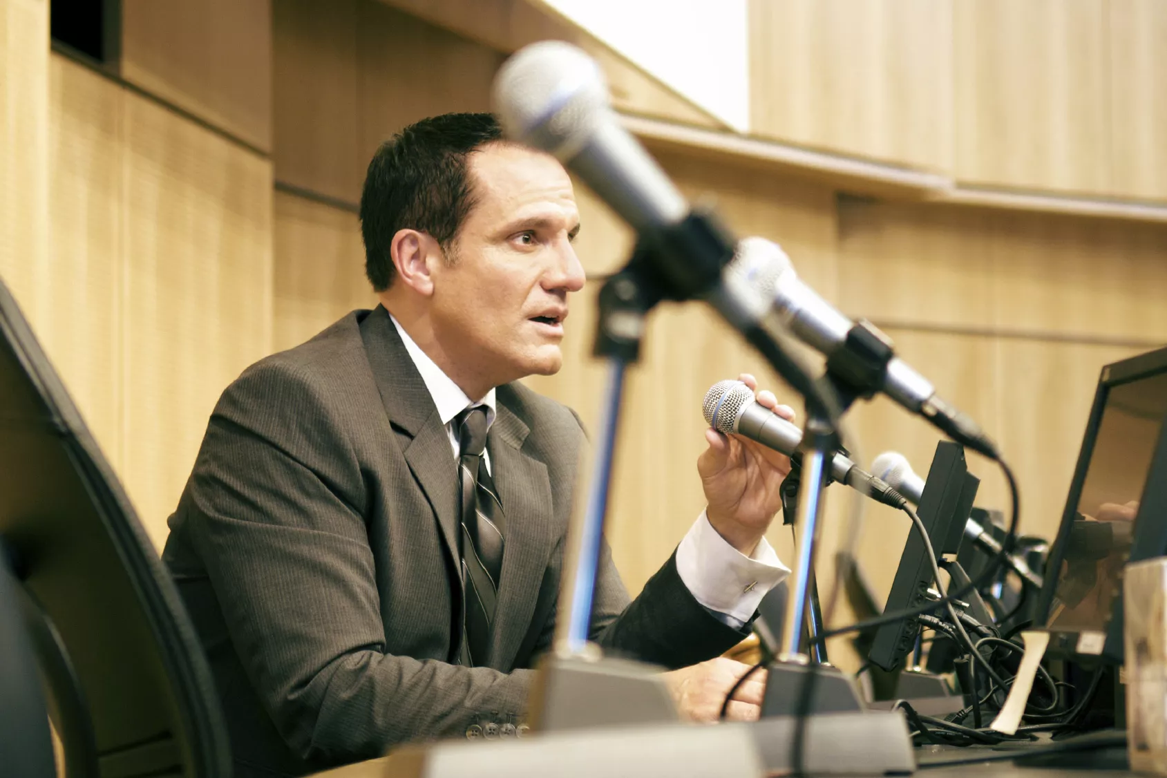 A speaker in a suit holding a microphone during a conference or meeting. The image features multiple microphones in the foreground and a professional setting in the background, emphasizing a formal and communicative atmosphere. 